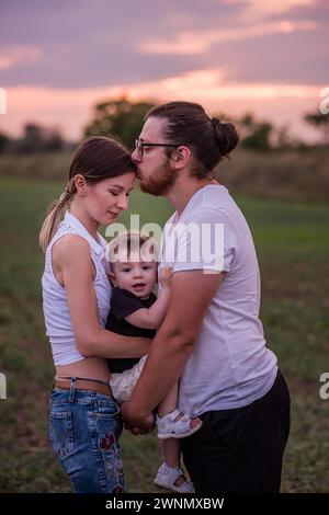 Vielfalt stilvoller Vater küsst Mütter in die Stirn, während er den kleinen Sohn hält, Konzept der schönen Familienverbindung vor dem pulsierenden Sonnenuntergang. Familienbande, Stockfoto