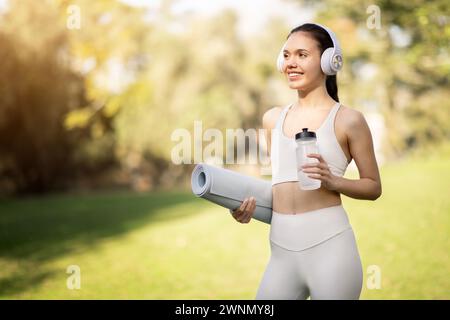 Lächelnde Frau mit Kopfhörern und Yogamatte genießt einen sonnigen Tag im Park, bereit für ein Wellness-Workout Stockfoto