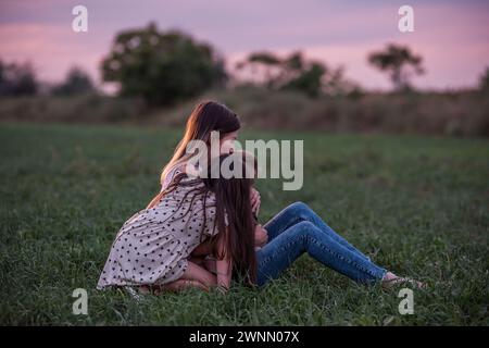 Mutter umarmt Sohn und Tochter, sitzt auf Gras, während die Dämmerung sich einsetzt. Moment der stillen Zuneigung, Verbindung. Ruhige Familienzeit in der Abenddämmerung. W Stockfoto