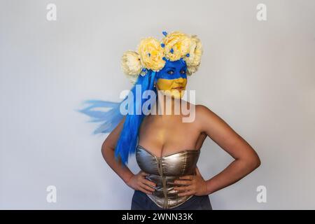 Schöne kreative junge Frau, die lächelt, während sie die Kamera auf isoliertem Hintergrund betrachtet. Mädchen in hellem Korsett gekleidet, blaue Haare, gelbe blaue Gesichtskunst, Stockfoto