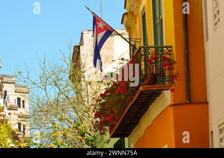 Kuba-Flagge in einem historischen orangen Gebäude, ein Balkon mit Metallrahmen und rotem Blumengarten, Blick auf die Baumspitze und einen blauen sonnigen Himmel. Stockfoto