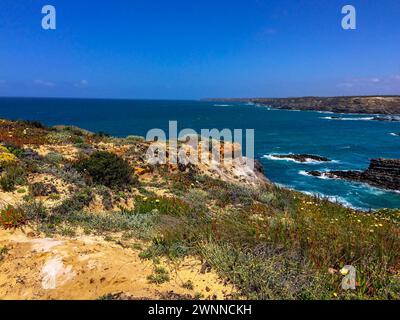 Eine Küstenlandschaft mit felsigen Klippen, Grün und einem klaren blauen Himmel. Die Meereswellen krachen gegen die Felsen. Stockfoto