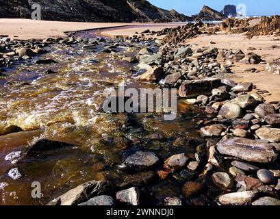 Klares Wasser rauscht über Steine und führt zu einem ruhigen Strand mit Klippen in der Ferne. Stockfoto