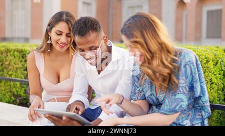 Lächelnde Freunde sitzen auf der Straße, während sie ein Tablet verwenden Stockfoto