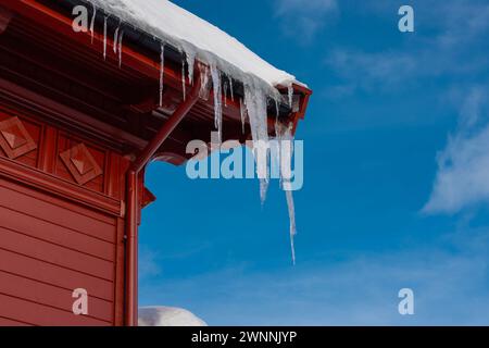 Lange gefährliche Eiszapfen hängen von einem Dach eines roten Holzhauses. Typisches Haus aus einer norwegischen Region, bedeckt mit Schnee und Eiszapfen. Stockfoto