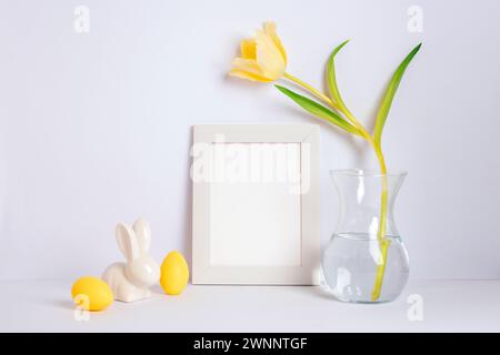 Leerer Bilderrahmen, gelbe Tulpe in Vase und Osterdekorationen auf weißem Hintergrund. Osterkonzept. Stockfoto