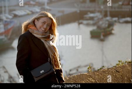 Porträt einer blonden Frau, die lächelt, sie sieht sehr glücklich aus, sie trägt einen dunklen Mantel und einen braun-weißen Schal, es ist ein sonniger Wintertag und in der Stockfoto