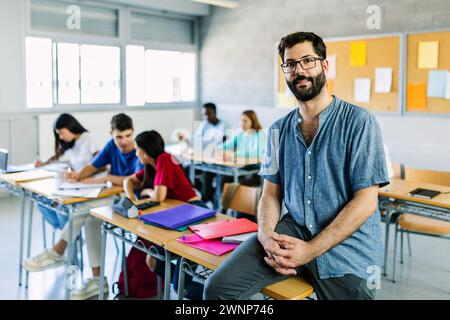 Porträt eines jungen bärtigen männlichen Lehrers, der am High School-Tisch sitzt Stockfoto