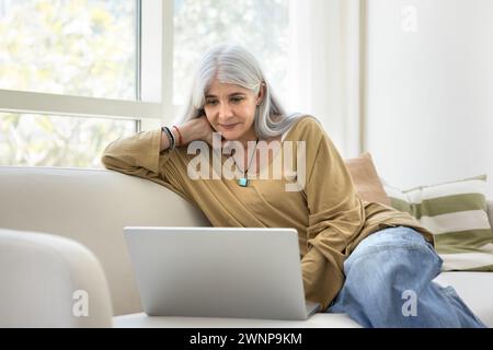 Positive grauhaarige ältere hispanische Frau, die sich mit dem Laptop entspannt Stockfoto