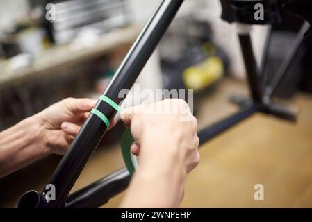 Unerkennbarer Mann, der einen Fahrradrahmen mit Abdeckband für ein individuelles Maldesign in seiner Fahrradwerkstatt vorbereitet, ein handgefertigter kreativer Prozess. Stockfoto