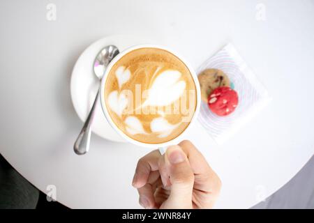 Cappuccino in einer weißen Tasse auf einem weißen Tisch mit zwei Mini-Keksen, Schokoladenchips und rotem Samt. Stockfoto