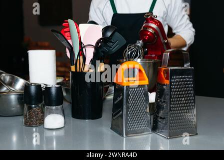 Eine Vielzahl von Küchenutensilien und Gadgets, die ordentlich auf einer Arbeitsplatte angeordnet sind, mit einem Koch im Hintergrund. Stockfoto