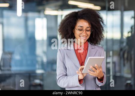 Eine lächelnde junge afroamerikanische Geschäftsfrau steht in einem Anzug im Büro und lächelt ein Tablet in ihren Händen an. Stockfoto