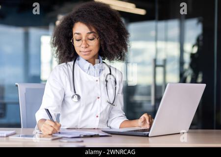 Ernsthafte junge afroamerikanische Ärztin sitzt an einem Schreibtisch in einem Krankenhaus und konzentriert sich auf Notizen in Dokumenten. Stockfoto