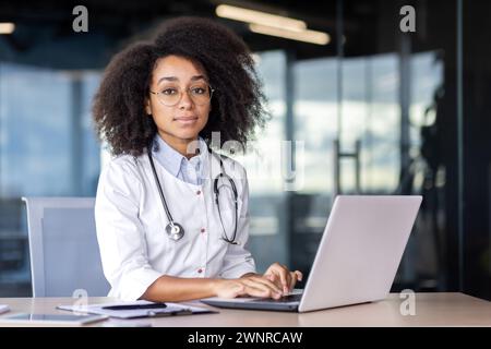 Porträt einer ernsthaften, selbstbewussten Ärztin, die in einem Krankenhausbüro an einem Schreibtisch sitzt, an einem Laptop arbeitet und die Kamera betrachtet. Stockfoto