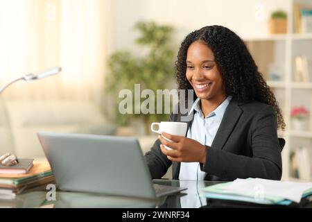 Glücklicher schwarzer Tele-Mitarbeiter, der online mit einem Laptop arbeitet und zu Hause Kaffee trinkt Stockfoto