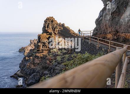 Frau, die am Ende eines Weges zwischen Felsen auf einer Klippe steht, vor einem riesigen Felsen und dem Meer, in La Palma, Kanarische Insel Stockfoto