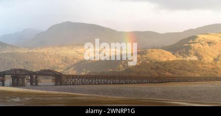 Ein Sonnenstrahl bricht durch die Gewitterwolken und bildet einen Regenbogen über der Barmouth Bridge (Pont Abermaw) Gwynedd Wales UK. März 2024 Stockfoto