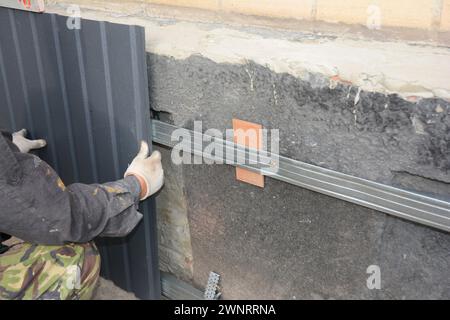 Schließen Sie die Hände des Bauherstellers, die Metallbleche installieren, um das alte Hausfundament vor Regen abzudichten und zu renovieren. Stockfoto