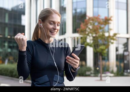 Eine fröhliche Frau feiert einen Moment des Sieges oder der guten Nachrichten im Freien, indem sie Musik über Kopfhörer von ihrem Smartphone hört. Die Kulisse zeigt urbanes Grün und moderne Architektur, die ein Gefühl der Vereinbarkeit von Beruf und Privatleben vermittelt. Stockfoto