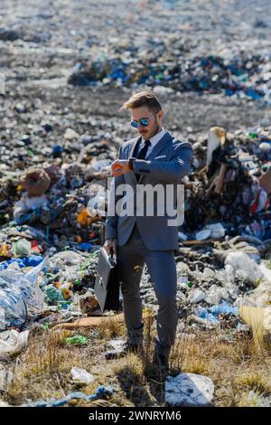 Geschäftsmann, der auf die Uhr blickt, die Zeit auf der Mülldeponie sieht, großer Müllhaufen. Konsum versus Verschmutzungskonzept. Soziale Netzwerke im Unternehmen Stockfoto