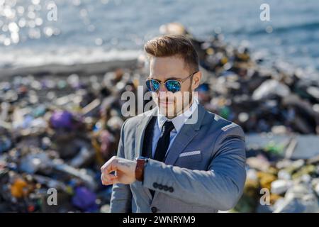 Geschäftsmann, der auf die Uhr blickt, die Zeit überprüft, die auf einem Müllhaufen am Strand steht. Konsum versus Verschmutzungskonzept. Soziale Verantwortung von Unternehmen Stockfoto