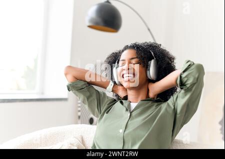 Glückselig und entspannt dehnt sich die Frau, während sie Kopfhörer trägt, und macht eine Pause von der Arbeit oder dem Alltag in ihrem gemütlichen Zuhause. Die Szene ist eine perfekte Mischung aus Komfort und modernem Wohnen Stockfoto