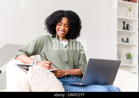 Eine afroamerikanische Frau mit Inhalten betreibt Multitasking, schreibt in einem Notebook und verwaltet gleichzeitig Aufgaben auf ihrem Laptop in ihrem ruhigen Heimbüro Stockfoto