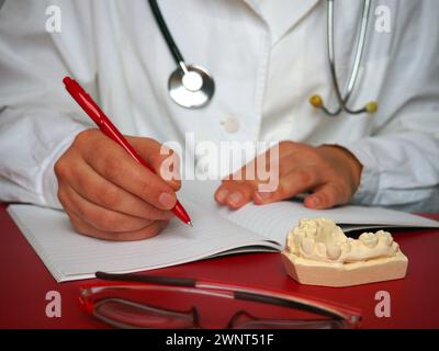 Der Arzt schreibt etwas in ein Notizbuch oder Notizbuch. Weißes medizinisches Kleid. Brille und ein Gipsknochen stehen auf dem roten Tisch. Ein Stethoskop hängt Stockfoto