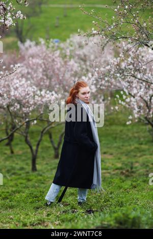 Auf den Frühling wartet ein Mädchen in einem schwarzen langen Mantel durch einen blühenden Garten Stockfoto