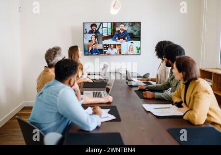 Das Team hat ein virtuelles Meeting mit einem Bildschirm durchgeführt, auf dem sowohl Remote-Teilnehmer als auch persönliche Kollegen nahtlos interagieren, und die Integration moderner Technologien in kollaborative Arbeitsbereiche Stockfoto