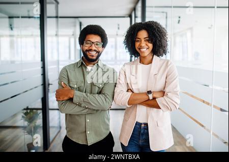 Zwei lächelnde Kollegen, ein Mann und eine Frau, stehen selbstbewusst in einem modernen Bürobereich, mit überkreuzten Armen und symbolisieren starke Teamarbeit und Führungsstärke in einem dynamischen und professionellen Umfeld. Stockfoto