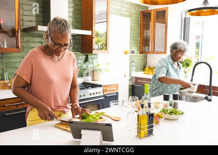 Eine ranghohe Afroamerikanerin hackt Gemüse, während eine ranghohe Frau Produkte wäscht Stockfoto