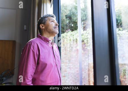 Ein älterer birassischer Mann in einem rosa Hemd sieht kontemplativ aus einem Fenster mit Kopierraum aus Stockfoto