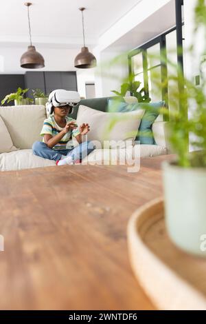 Ein afroamerikanischer Junge mit VR-Headset sitzt auf einer Couch, in ein virtuelles Spiel eingetaucht, mit Kopierraum Stockfoto