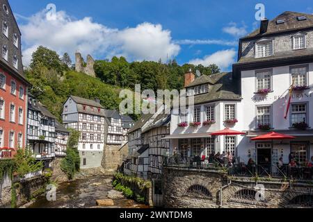 Malerisches europäisches Dorf (Monschau, Deutschland) mit Fachwerkhäusern und einer blumengesäumten Brücke über einen Bach mit grünen Hügeln im Hintergrund Stockfoto