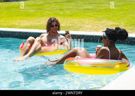 Ein vielfältiges Paar junger Kaukasier und Afroamerikaner entspannen sich auf bunten Schwimmbädern in einem Pool Stockfoto