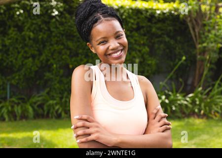 Die junge afroamerikanische Frau lächelt hell, die Arme in einem lässigen pfirsichfarbenen Oberteil gekreuzt Stockfoto