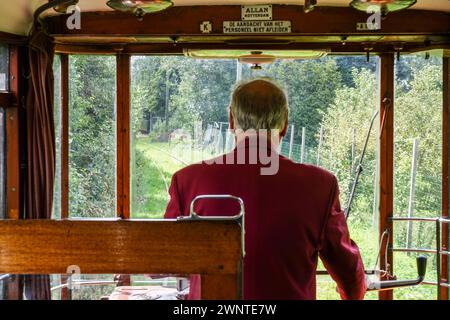 Straßenbahnführer in roter Jacke mit Blick aus der Kabine einer alten Straßenbahn, die malerische Eisenbahnstrecke und Bäume in Amsterdam, Niederlande, beobachtet Stockfoto