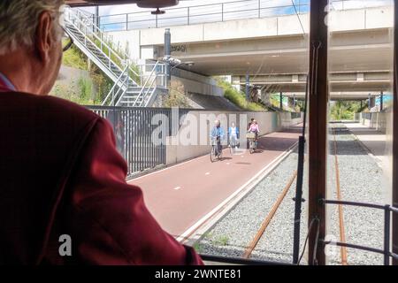 Blick vom Straßenbahnfenster hinter dem Schaffner auf Menschen, die an einem sonnigen Tag in Amsterdam auf einem Stadtweg unter einer Brücke spazieren und radeln Stockfoto