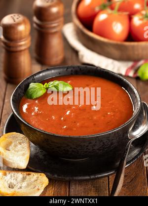 Schüssel Tomatensuppe garniert mit Basilikumblättern und gerösteten Baguetteschnitten, frische Tomaten im Hintergrund auf Holztisch Stockfoto