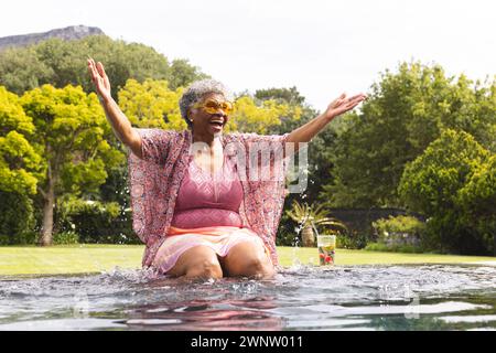 Eine ältere birassische Frau genießt einen erfrischenden Moment in einem Pool, ihre Arme sind vor Freude weit gespreizt Stockfoto