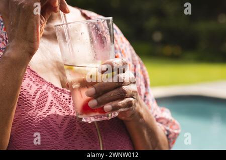 Eine ältere Frau mit birassischer Herkunft genießt einen erfrischenden Drink am Pool, ihre Hand hält sanft ein Glas Stockfoto