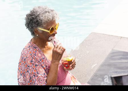 Eine ältere Frau mit Birasität genießt einen Drink am Pool, trägt eine gelbe Sonnenbrille und ein gemustertes Oberteil Stockfoto