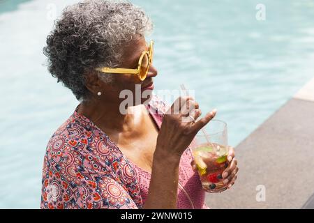 Eine ältere Frau mit einer gelben Sonnenbrille genießt einen Drink am Pool Stockfoto