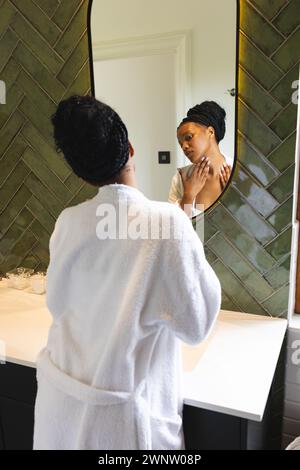 Eine junge afroamerikanische Frau in einem weißen Gewand untersucht ihr Spiegelbild im Badezimmer Stockfoto