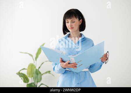 Professionelle Geschäftsfrau im formellen blauen Hemd prüft Papierdokumente in der Mappe steht auf weißem Wandhintergrund mit großer grüner Pflanze. Rechtsanwalt, Rechtsberater, Büroassistent oder Unternehmensleitung. Stockfoto