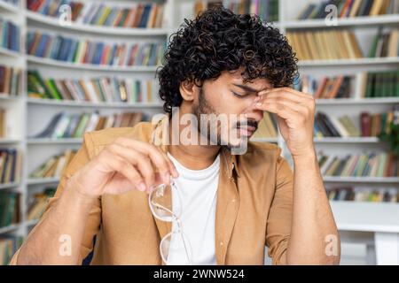 Ein junger Mann mit lockigen Haaren wirkt gestresst und hat Schmerzen im Kopf. Er zeigt Anzeichen von Kopfschmerzen. Er hat seine Brille abgenommen, vielleicht nachdem er lange Stunden gelesen oder studiert hat. Stockfoto