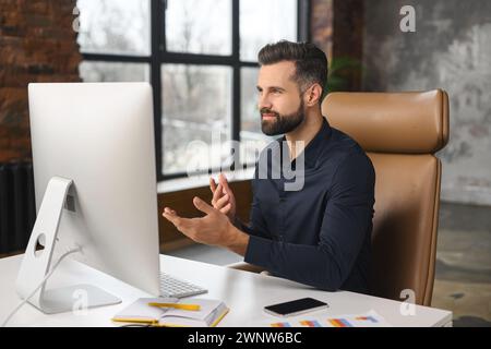 Junger bärtiger, selbstbewusster erfolgreicher Mann in Business-Freizeitkleidung, der während eines Videoanrufs spricht, ist ein erfahrener Bewerber bereit für ein Online-Interview bei einem Videoanruf, sitzt am Schreibtisch Stockfoto