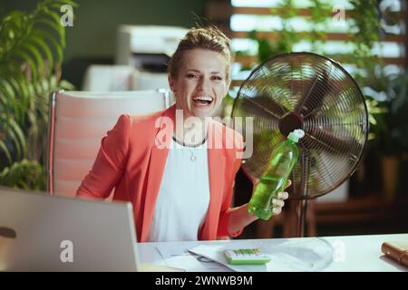 Nachhaltiger Arbeitsplatz. Porträt einer glücklichen modernen 40-jährigen Geschäftsfrau bei der Arbeit in roter Jacke mit Wasserflasche, elektrischem Ventilator und Laptop. Stockfoto